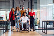 © .shock - A diverse group of young business people walking a corridor in the glass-enclosed office of a modern startup, including a person in a wheelchair and a woman wearing a hijab