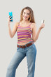 Smiling young woman displays mobile phone and credit card while browsing internet web page or online store, utilizing smartphone for shopping and online payments. Isolated on gray background