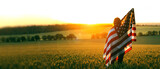 Fototapeta  - Independence Day. Beautiful girl with the American flag in a wheat field at sunset. 4th of July. Patriotic holiday, american day.