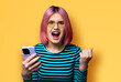 Excited surprised shocked astonished very happy pink woman wear braces sunglasses eye glass spectacles open mouth hold cell phone cellular smartphone cellphone isolated against yellow background.