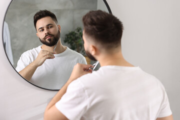 Canvas Print - Handsome young man trimming beard with scissors near mirror in bathroom