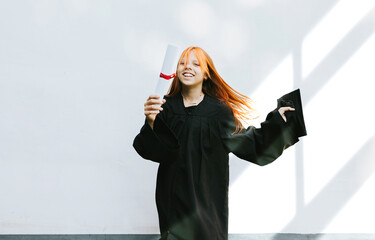 Wall Mural - teenage girl in clothes of a graduate coat and cap celebrates high school or  junior year graduation on background of white wall with shadows and with diploma in hands, education and no school concept