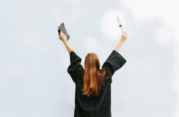 Wall Mural - teenage girl in clothes of a graduate coat and cap celebrates high school or  junior year graduation on background of white wall with shadows and with diploma in hands, education and no school concept