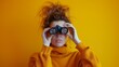 © OMD - Young woman in an orange sweater using binoculars, with a vibrant yellow background.