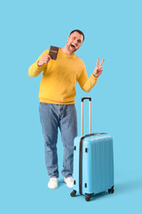 Wall Mural - Male tourist with suitcase and passport showing victory gesture on blue background