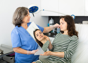 Wall Mural - Female client looking at mirror while qualified beautician examining her face skin after procedure in medical cosmetology office.