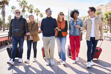 Wall Mural - Group of smiling mature people walking carefree with their luggage enjoy vacations on street of Barcelona city. Diverse happy middle-aged friends celebrating leisure time on spring sunny day