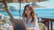© BOONJUNG - A female wearing white blouse is smiling and typing on a laptop on a seaside terrace