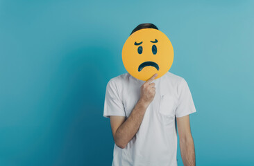 Wall Mural - A man holding an emoji with a sad face on it, wearing a white t-shirt against a light blue background