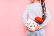 Little girl holding a drawn card and a flower behind her back for Father's Day holiday in front of a pink background