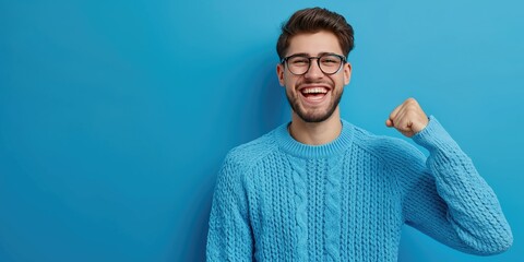 Wall Mural - A young handsome man wearing a blue sweater and glasses on background with a smiling face doing a winner gesture holding his fist up celebrating success or happy positive expression of good news,