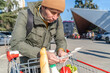 Man with a grocery cart outdoors, reviewing a shopping list, with fresh produce visible in the cart.