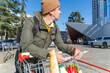 Man with a grocery cart outdoors, reviewing a shopping list, with fresh produce visible in the cart.