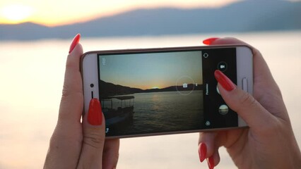 Detail view on female hands holding smartphone and making photo of scenic marine landscape at sunset time. Girl using phone to get beautiful pictures of seascape. Blurred background. Isolated shot