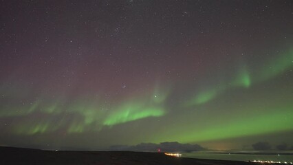 Wall Mural - Mesmerizing show of northern lights in the night sky above the sea. A small town stands on the coast illuminated by the lights. A timelapse video.