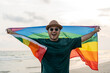 Young asian man with pride movement LGBT holding rainbow flag raise up for freedom. Demonstrate rights LGBTQ celebration pride Month Gay Pride Symbol. Standing on the sand sea beach during sunset