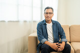 Fototapeta Sawanna - Handsome asian mature old man sitting on sofa couch in living room at home. Happy Portrait of cheerful smiling senior asian man relax in the house. Mature People and lifestyle