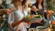 © Nijat - Happy young woman serving festive table with vegetable salad in bowl while standing against her friend bringing other snacks and appetizers