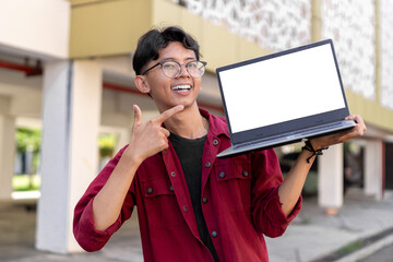 Young Asian man wearing red shirt smiling and showing white laptop screen for advertisement. Concept of people lifestyle. Happy man holding blank laptop screen with blurred background.