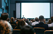 © Kien - an audience from the front and side views, with people sitting in chairs watching someone on stage presenting to them. A white screen for presentations is on a big wall behind the conference room