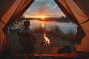 Wall Mural - A serene scene of a man seated outside a tent, gazing at a tranquil river as the sun sets, evoking calm and adventure