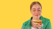 happy woman eating and looking at sandwich, hamburger on yellow background with copy space  unhealthy lifestyle