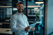 © wolfhound911 - A smiling, busy young Caucasian business man entrepreneur is standing in his office at work, immersed in a tablet. The happy male professional executive manager is using the tablet computer to review