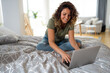 Young content woman using laptop for online work while sitting on the bed at home.