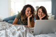 Two cheerful women lying on the bed in bedroom and watching movies on laptop.