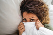Sad exhausted woman looking at camera wrapped up in a blanket up to her nose due to illness and cold or negative emotions.