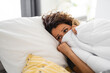 Sad young woman covered in duvet up to her nose lying in cozy bed. She is suffering from heat temperature, feeling cold and flu.