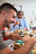 Vertical. Side view cheerful Caucasian man holding cutlery eating food sitting at table with his family unfocused in background. Happy people gathered to enjoy delicious meal in celebration at home