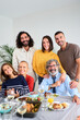 Vertical. Portrait happy Caucasian family posing smiling looking at camera hugging indoors. Multi-generational joyful people gathered around table full of food for celebrating meal together at home