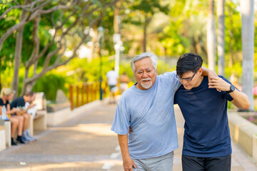 Elderly man suffering knee pain during jogging exercise with his son at park. A man support father with knee injury walking on jogging track. Family relationship and senior people health care concept.