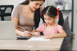 Mother helps daughter doing homework sitting at table writing in paper notebook right answer