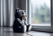 A black and white photo of a teddy bear sitting alone next to a window on a rainy day