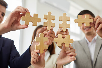 Happy smiling group of business people assembling wooden puzzle standing in office. Company employees join jigsaw pieces finding solution. Cooperation, teamwork, help and support concept.