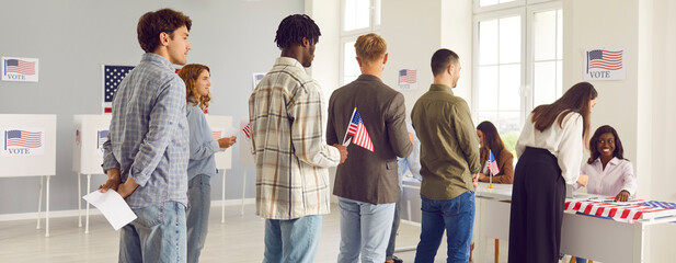 Wall Mural - Large group of diverse people registering at polling station holding American flags in hands on election day. American voters standing in queue at vote center among citizens of USA. Banner.