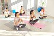 Group of young sporty women carelessly practice yoga sitting in lotus position in sunny studio. Focused individuals practicing yoga poses in peaceful studio, embodying tranquility and mindfulness.