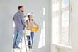 Young family couple doing renovations and painting walls inside house. Happy, joyful man and woman standing on ladder at home, holding bucket and rollers and looking at light walls and sunny windows