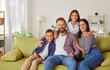 Happy young smiling family of four with two kids boy and girl sitting on the sofa at home and looking cheerful at camera. Portrait of parents with children. Family time and leisure concept.