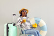 Funny woman who is expecting vacation is impressed by discounts on summer tours. Dark-skinned woman with cocktail, suitcase and inflatable ring sitting on chair with shocked expression on her face.