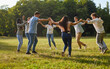 Group of happy diverse friends enjoying summer and having fun in beautiful sunny park. Eight cheerful young multiracial people holding hands and dancing round dance on green grassy lawn all together