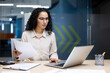 © Liubomir - Serious and pensive business woman behind paper work inside office, female financier worker thinks about contracts and reports with charts and graphs, hispanic successful woman uses laptop at work