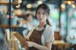 © darshika - Portrait of Asian girl waitress holding menu wearing apron and standing in coffee shop.