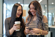 © 수동 김 - Two young women in stylish attire, smiling and holding coffee to go cups while looking at their phones together in an office setting
