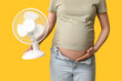 Pregnant young woman with electric fan on yellow background, closeup