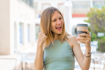 Young blonde woman holding a take away coffee at outdoors celebrating a victory