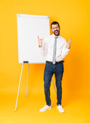 Wall Mural - Full-length shot of businessman giving a presentation on white board over isolated yellow background making rock gesture