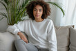 Mockup. Young woman wearing blank white crewneck sweatshirt. Young African American girl sitting on sofa in modern living room. Mock up template for sweatshirt design, print area for logo or design